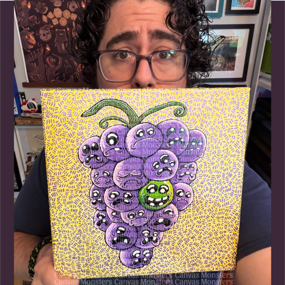 Mike, owner of Canvas Monsters, holding up a square canvas painting featuring a cluster of purple cartoon grapes with sad faces on a yellow patterned background. Mike is wearing glasses and a dark blue shirt, with framed artwork and shelves visible in the background.