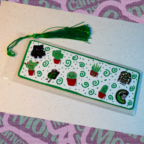 White paper bookmark decorated with mini green-themed Canvas Monsters stickers with a painted green border, in a plastic sleeve and a green tassel.