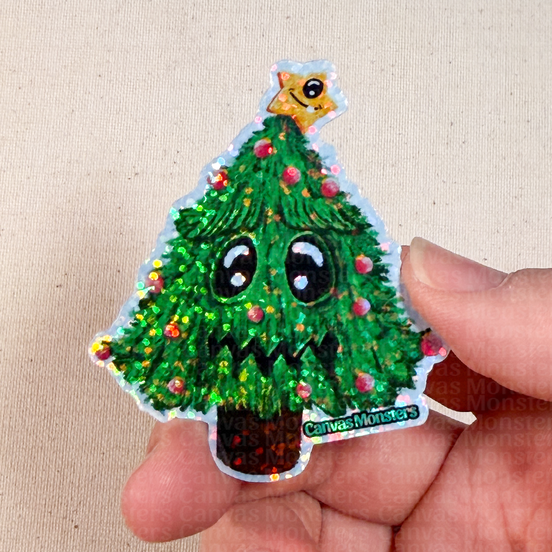 Hand holding a glitter sticker of a cartoon Christmas tree with a star on top against a beige background.
