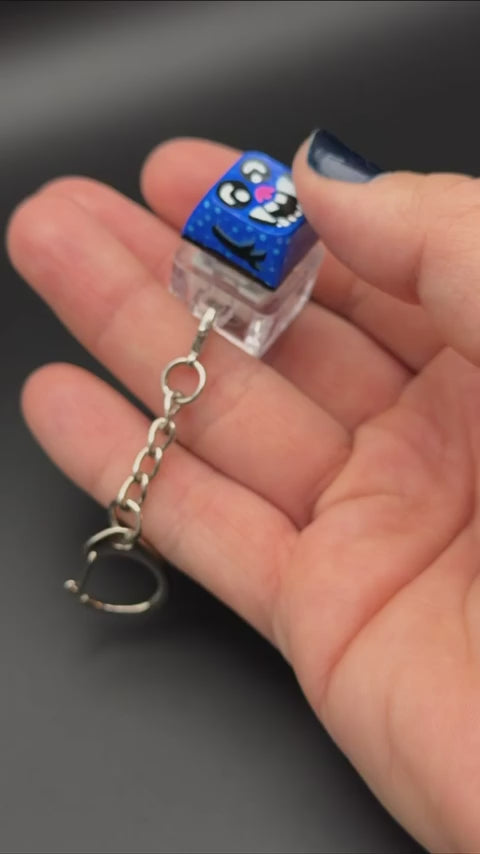 Video of a key cap keychain in action against a black background. Cap is painted with a blue monster character.