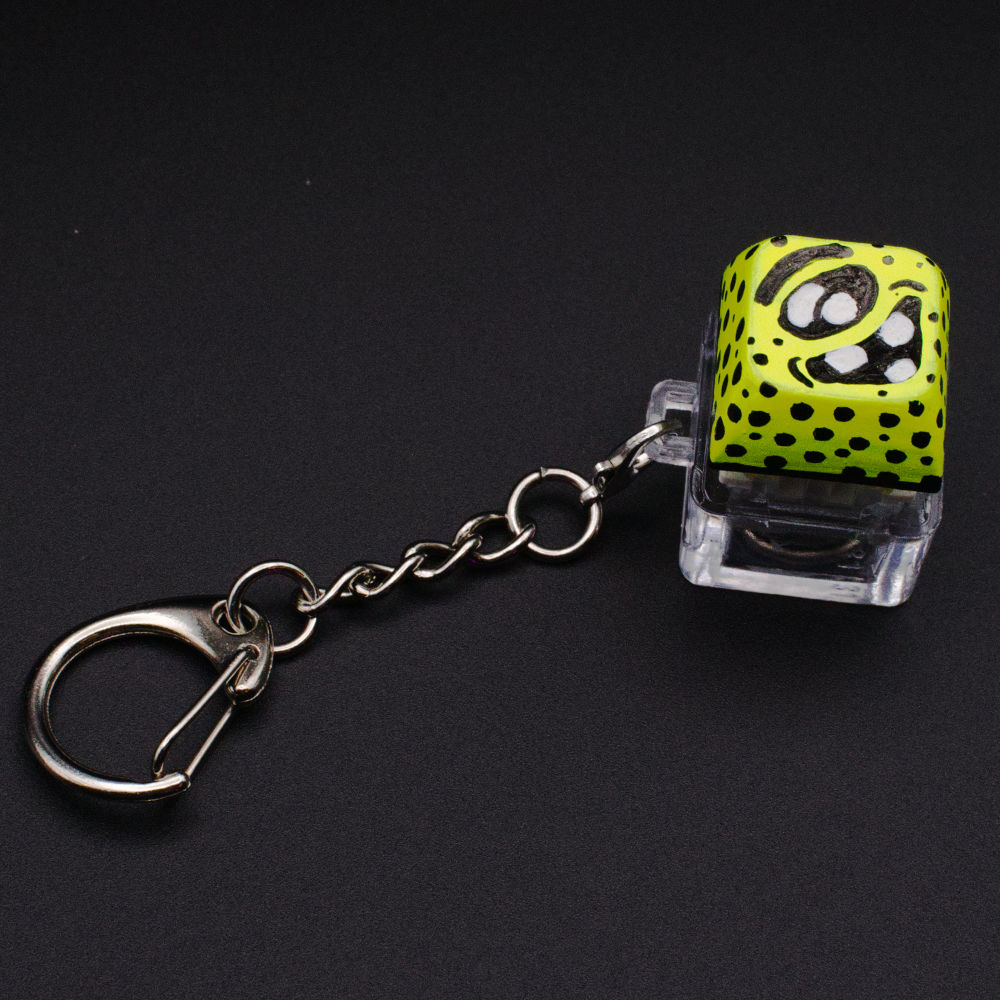 Photo of a key cap keychain on a black background. Cap is painted with a yellow monster character.