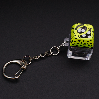 Photo of a key cap keychain on a black background. Cap is painted with a yellow monster character.