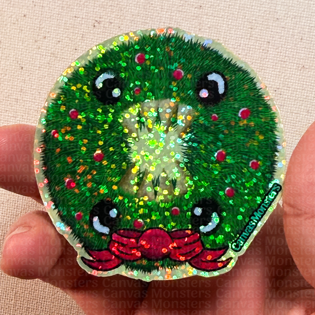 Glitter sticker of a cartoonish Holiday wreath with 4 eyes, 8 teeth, and a red holiday bow on a beige fabric background