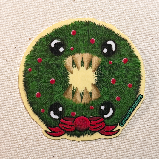 Glossy sticker of a cartoonish Holiday wreath with 4 eyes, 8 teeth, and a red holiday bow on a beige fabric background