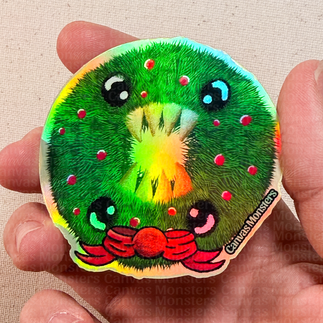 Holofoil sticker of a cartoonish Holiday wreath with 4 eyes, 8 teeth, and a red holiday bow on a beige fabric background